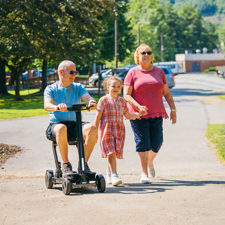 Man on a pride go go carbon mobility scooter with a young girl and woman walking alongside him on a sunny day.