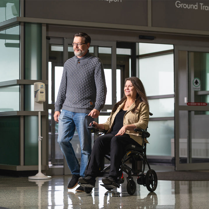 Man pushing a woman in a Black Pride Jazzy 27x  through a building entrance.