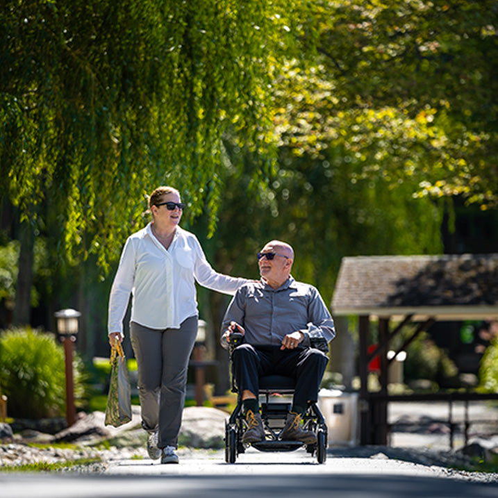 Woman pushing a man in a Pride Jazzy Ultra Light Power Chair through a park with greenery and a gazebo in the background.