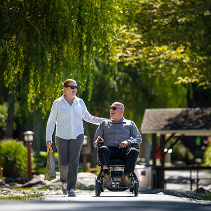Woman pushing a man in a Pride Jazzy Ultra Light Power Chair through a park with greenery and a gazebo in the background.