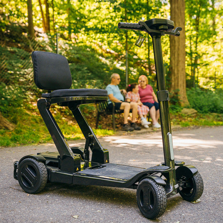Black pride go go carbon mobility scooter with a seat in a park setting, people sitting on a bench in the background.