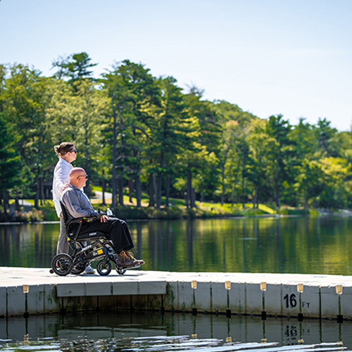 Person using a Pride Jazzy Ultra Light Power Chair by a lake with another person assisting, surrounded by trees.