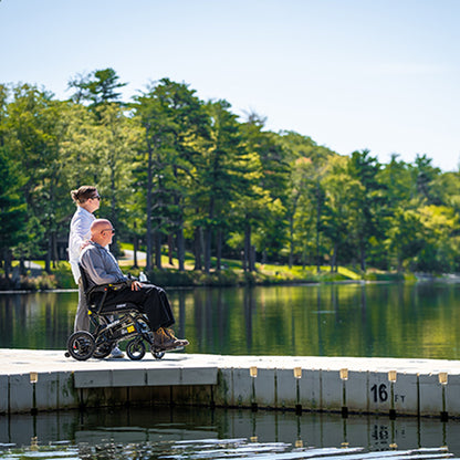 Person using a Pride Jazzy Ultra Light Power Chair by a lake with another person assisting, surrounded by trees.