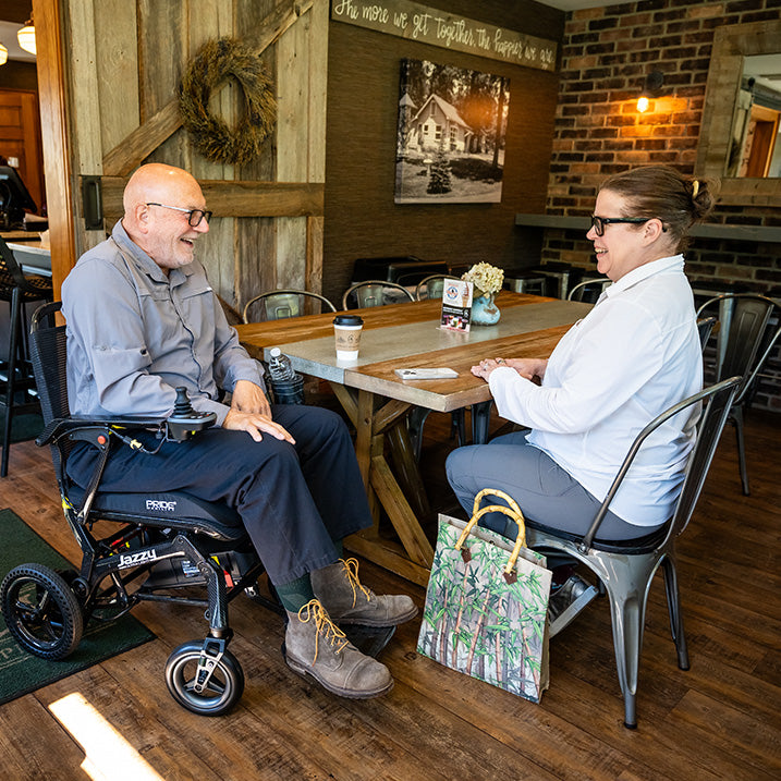 Two people sitting at a table in a cozy restaurant setting with a man in a pride jazzy ultra light wheelchair