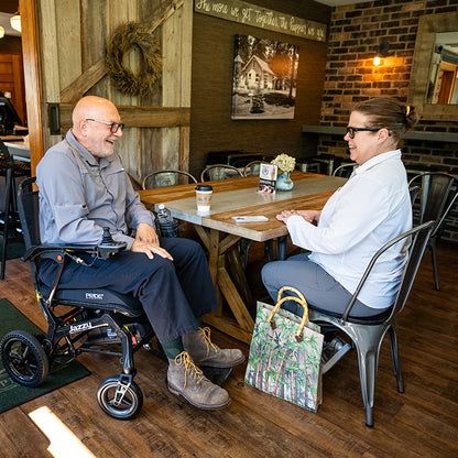 Two people sitting at a table in a cozy restaurant, one in a Pride Jazzy Ultra Light Power Chair.