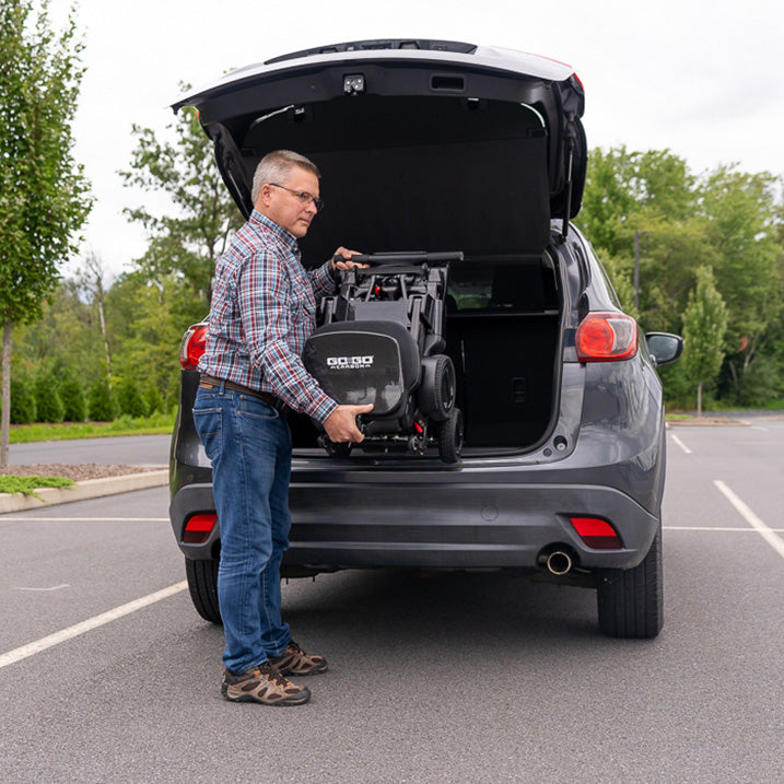 Man loading a  pride go go carbon mobility scooter into the trunk of a car in a parking lot.