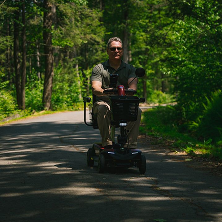 Man riding a Pride Go-Go Endurance AL+ scooter  on a paved path surrounded by trees