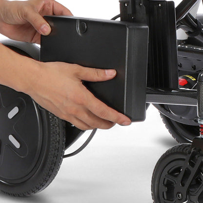 A close-up of a person's hands attaching a black rectangular battery pack to a black electric wheelchair or stroller frame with wheels visible.