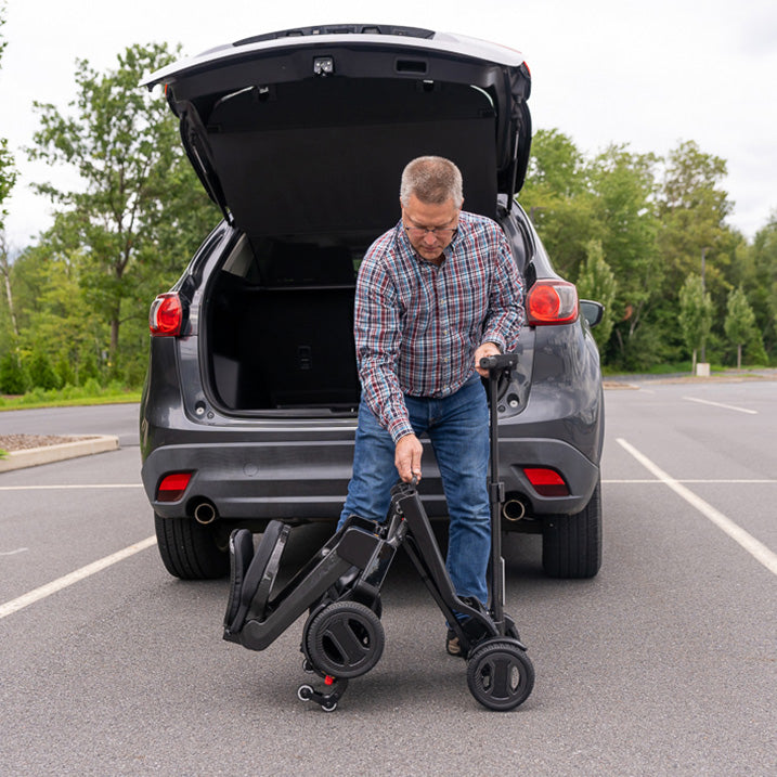 Man loading a  pride go go carbon scooter into the trunk of a car in a parking lot.