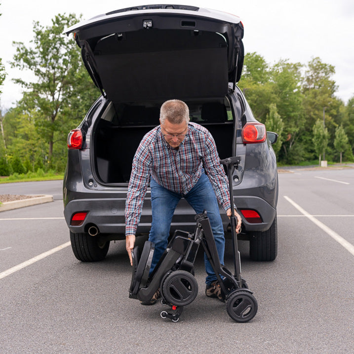 Man unloading a folded  pride go go carbon scooter from the trunk of a car in a parking lot.