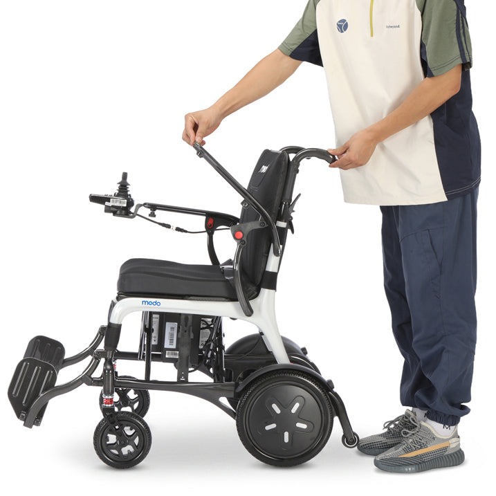 A person in a white and navy uniform pushes a white and black Modo Breeze Ultralightweight Folding Electric Wheelchair with a footrest on a clean white background.
