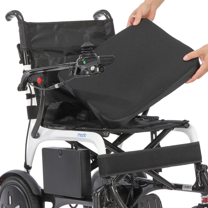 A person's hands are shown placing a black textured mesh backrest cushion onto Modo Breeze Ultralightweight Folding Electric Wheelchair, with the brand name 'mode' visible on the frame.