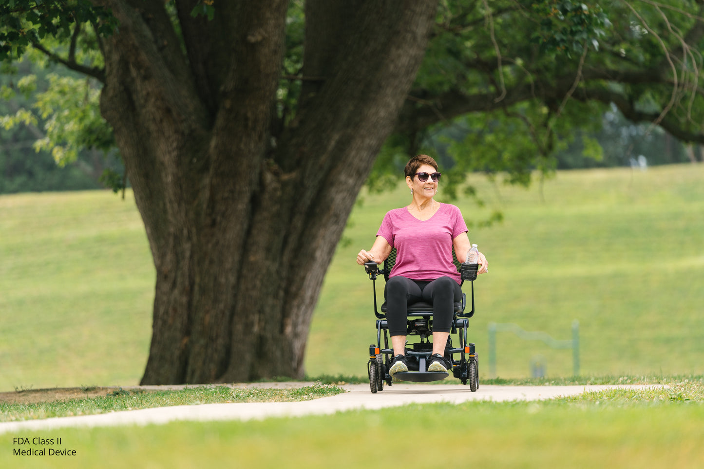 Woman using a mobility scooter in a park with a large tree and grassy area.