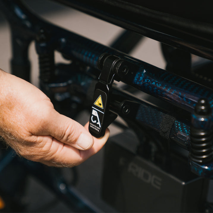 Hand holding a black lock with a yellow warning triangle on a vehicle's suspension system.