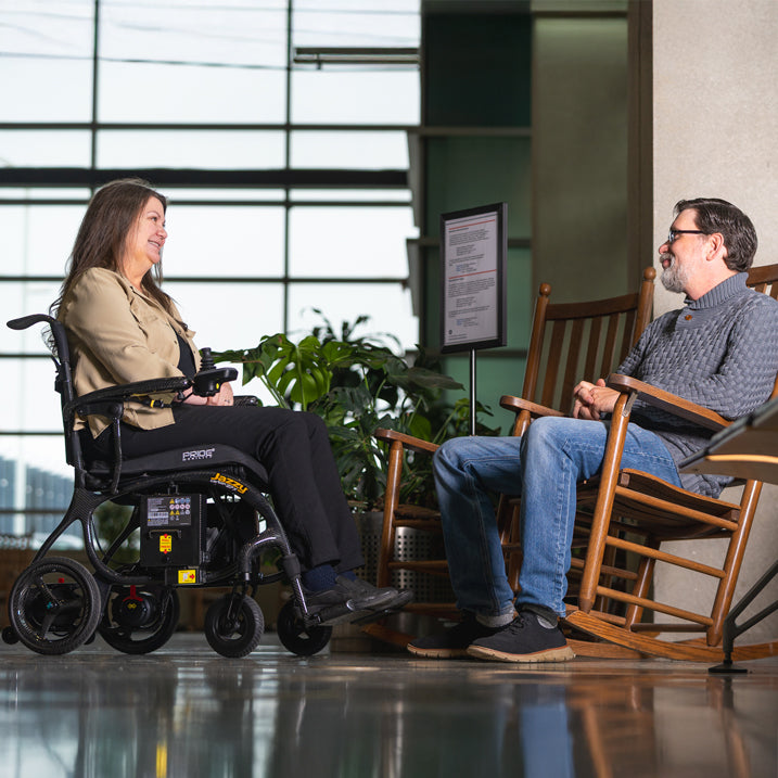 Person in a Black Pride Jazzy 27x  interacting with another person in a room with large windows and plants.