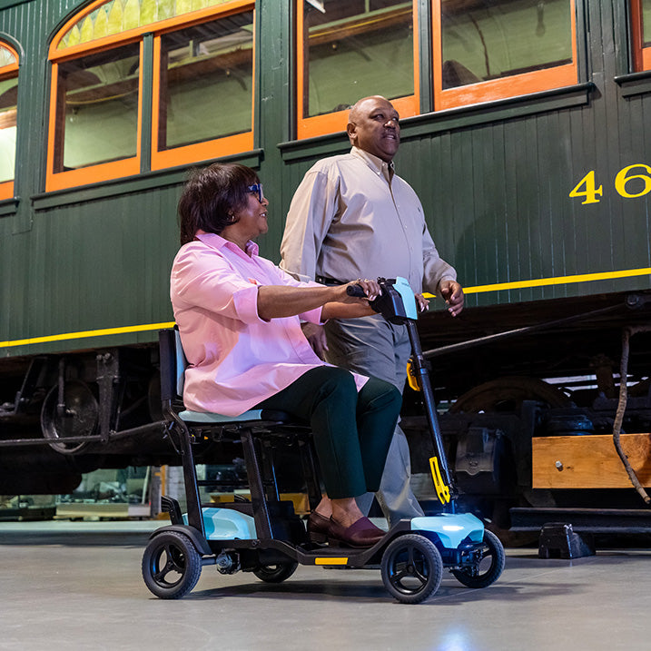 Woman using a pride go go super portable mobility scooter with a man standing behind her, in front of a vintage train car.