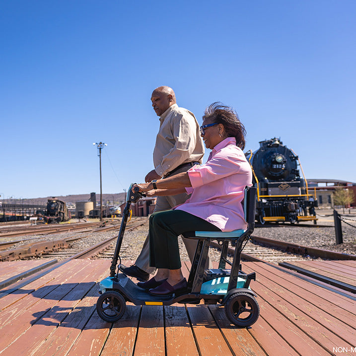 Person using a pride go go super portable mobility scooter with another person assisting them, set against a train background.