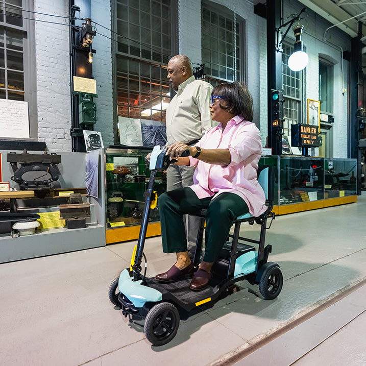 Person using a pride go go super portable mobility scooter in an urban setting with a building in the background