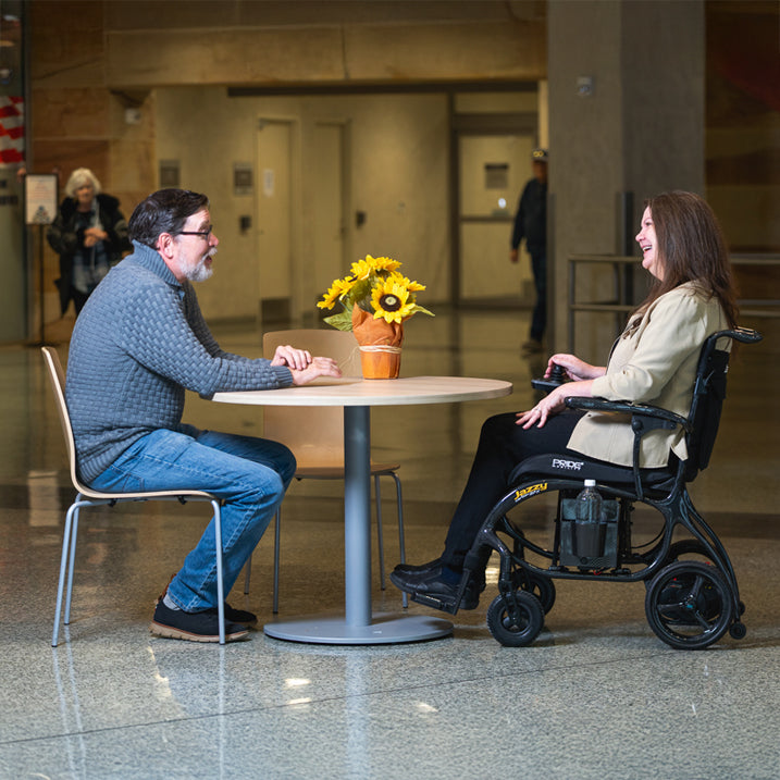 Two people, one in a wheelchair, sitting at a table in a public space.
