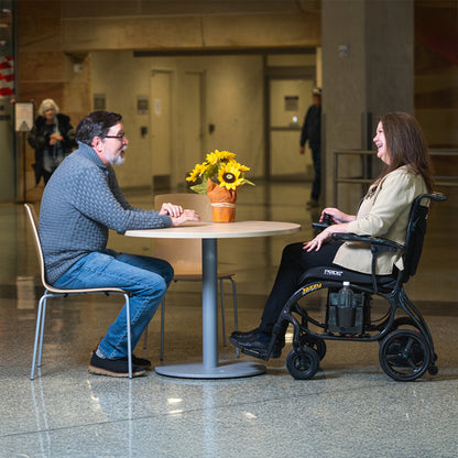 Two people, one in a wheelchair, sitting at a table in a public space.
