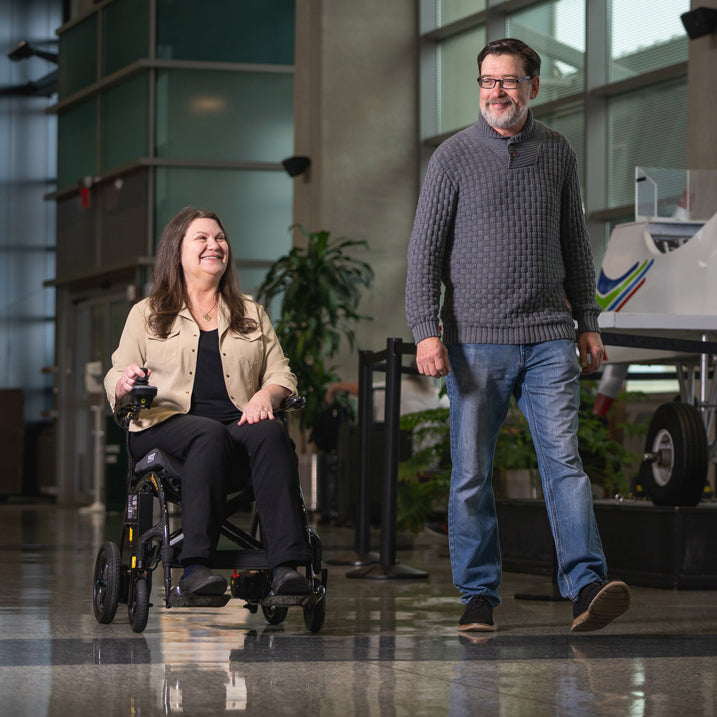 Woman in a wheelchair being pushed by a man in an indoor setting with large windows.