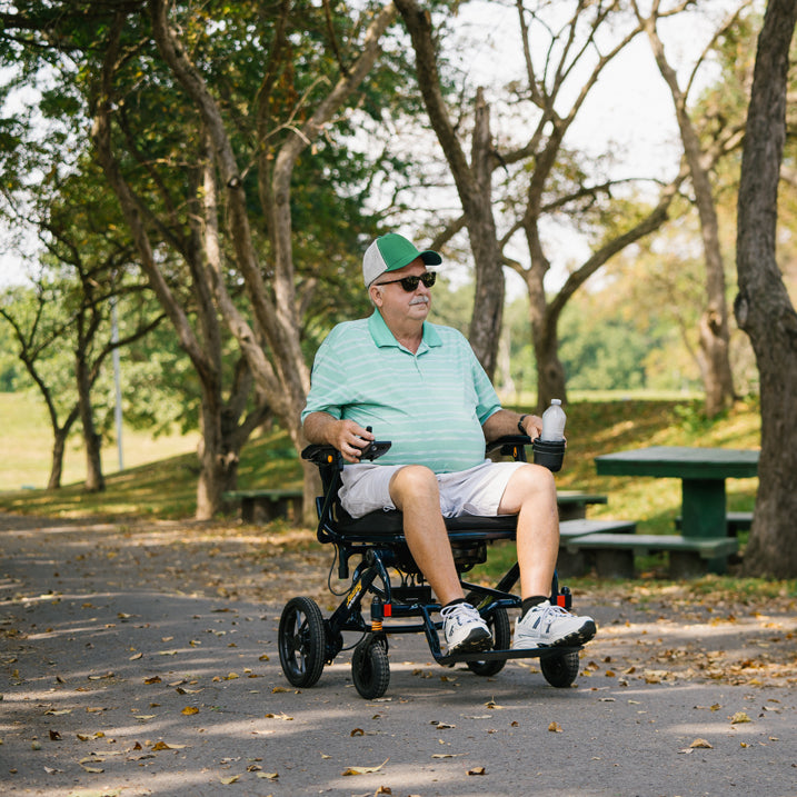 Man in a Jazzy carbon HD sitting on a path in a park with trees and a picnic table in the background.